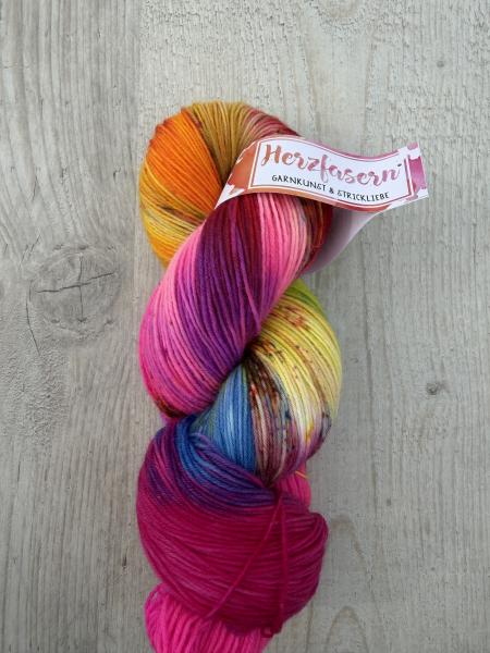 Handgefärbter Wollstrang in leuchtendem Pink mit Farbverläufen in Orange, Gelb, Blau und dunklen Sprenkeln auf hellem Holzuntergrund